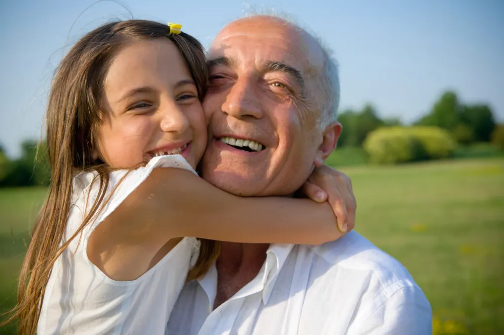 A girl hugs her grandfather, both laughing, on a sunny lawn